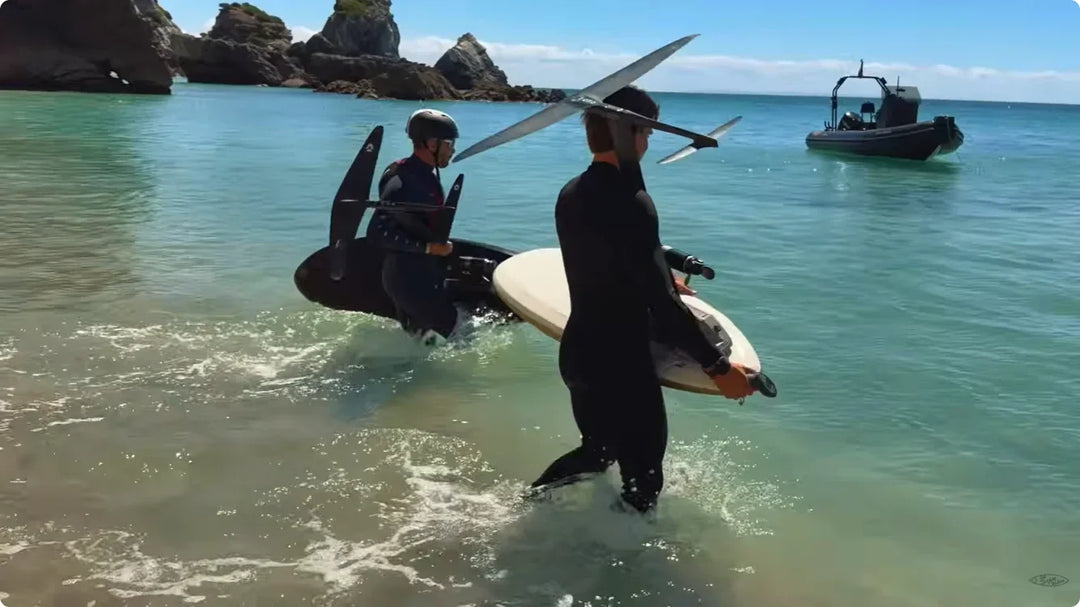 A rider glides on a FoilBoost hydrofoil board across clear turquoise water near anchored boats.
