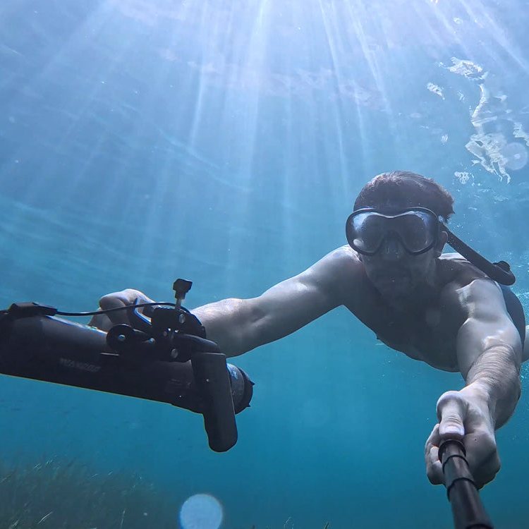 Person scuba diving underwater with a selfie stick, surrounded by sunlight filtering through the water.